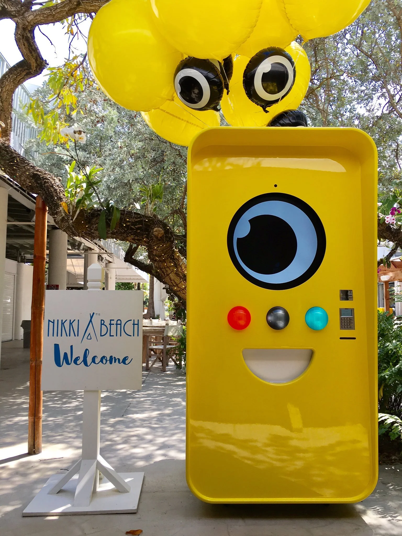 Yellow Snapbot vending machine with eye balloons beside a white NIKKI BEACH Welcome sign under a tree